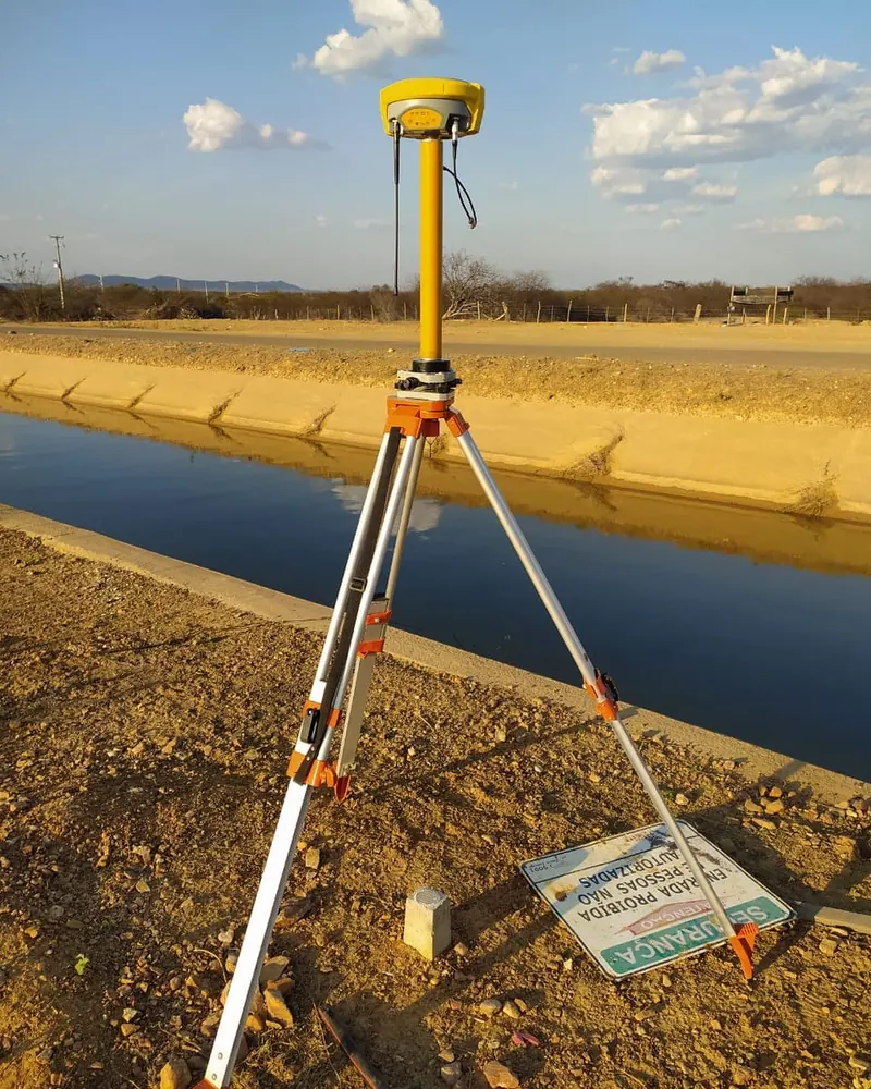 Receptor GNSS em tripé ao lado de um canal, durante levantamento de campo