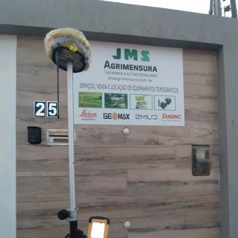 Receptor GNSS em frente à placa da JMS Agrimensura, com serviços, venda e locação de equipamentos topográficos